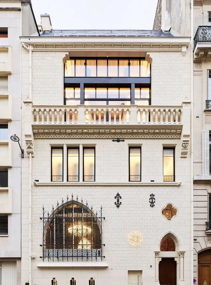 Façade d’hôtel particulier en pierre claire, grande baie en ogive grillagée et étage vitré en attique.