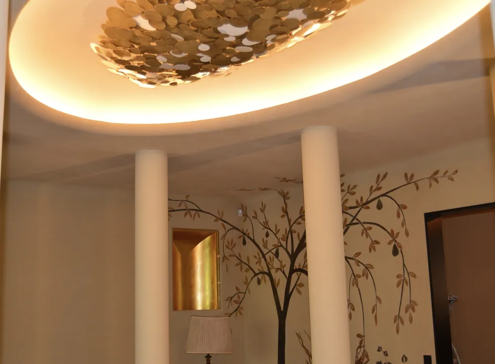 Hall intérieur avec deux colonnes blanches, plafond à éclairage indirect, niche dorée et arbre peint au mur.