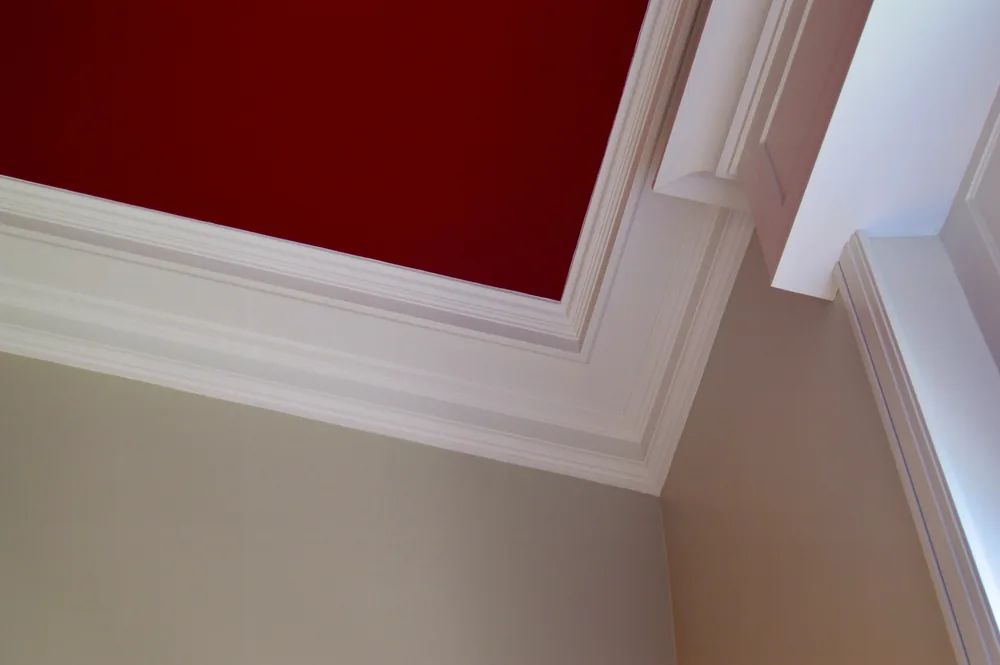 Angle de plafond peint en rouge profond, bordé de corniches et de moulures blanches, murs beige.