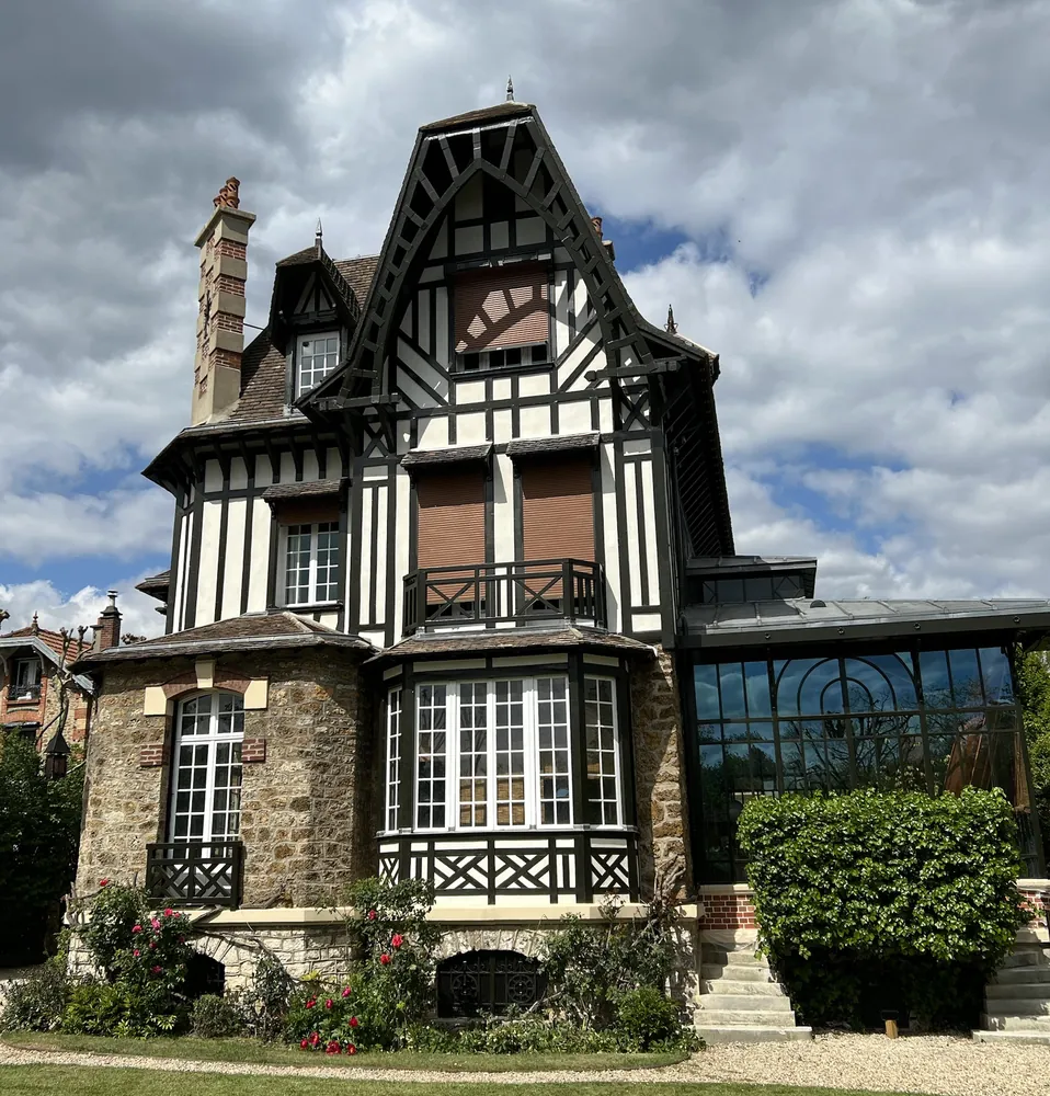 Villa à pans de bois noirs et remplissages blancs sur soubassement en meulière, avec une verrière à droite.