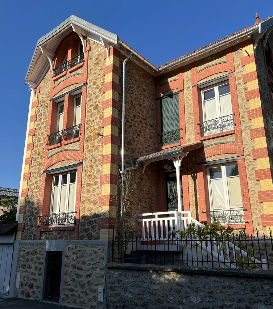 Villa en meulière aux chaînes d’angle en brique rouge et jaune, garde-corps en fonte et perron en bois blanc, sous un ciel bleu.